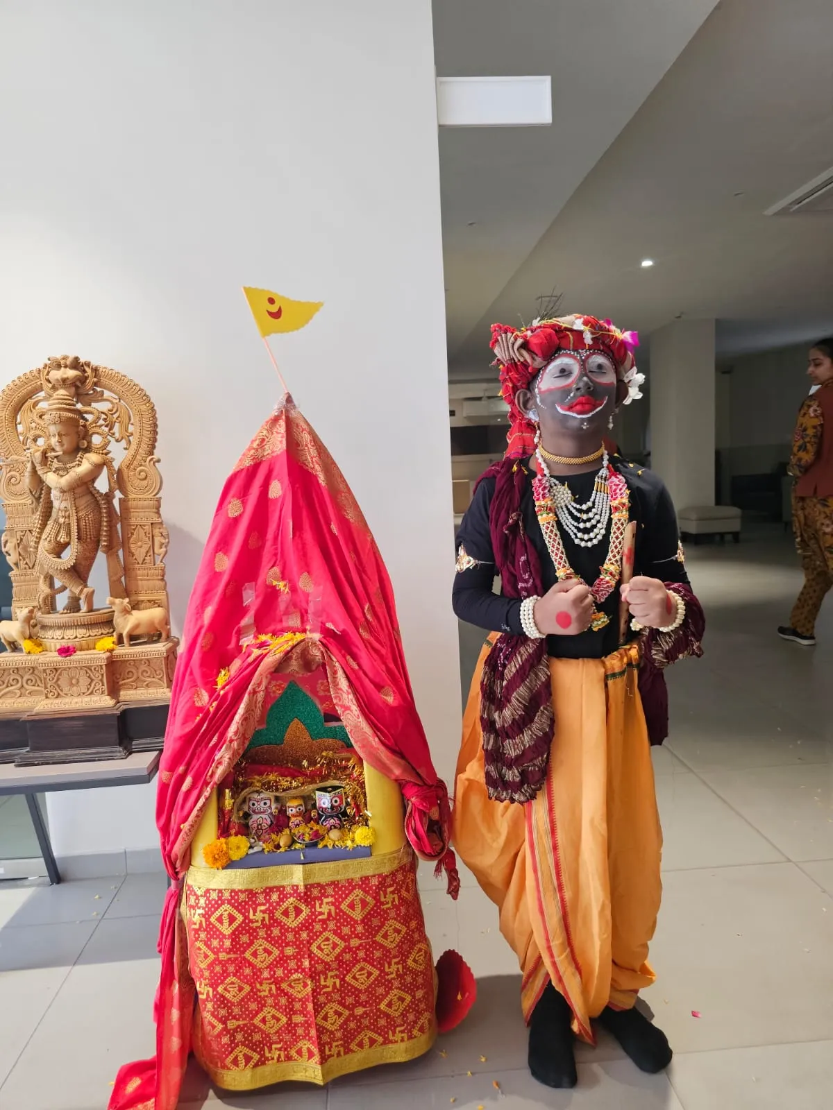 A child dressed in festive traditional attire, including a yellow dhoti and full dark face paint, standing proudly next to a small red decorated chariot at Shree Shakti School of Innovation (SSI).