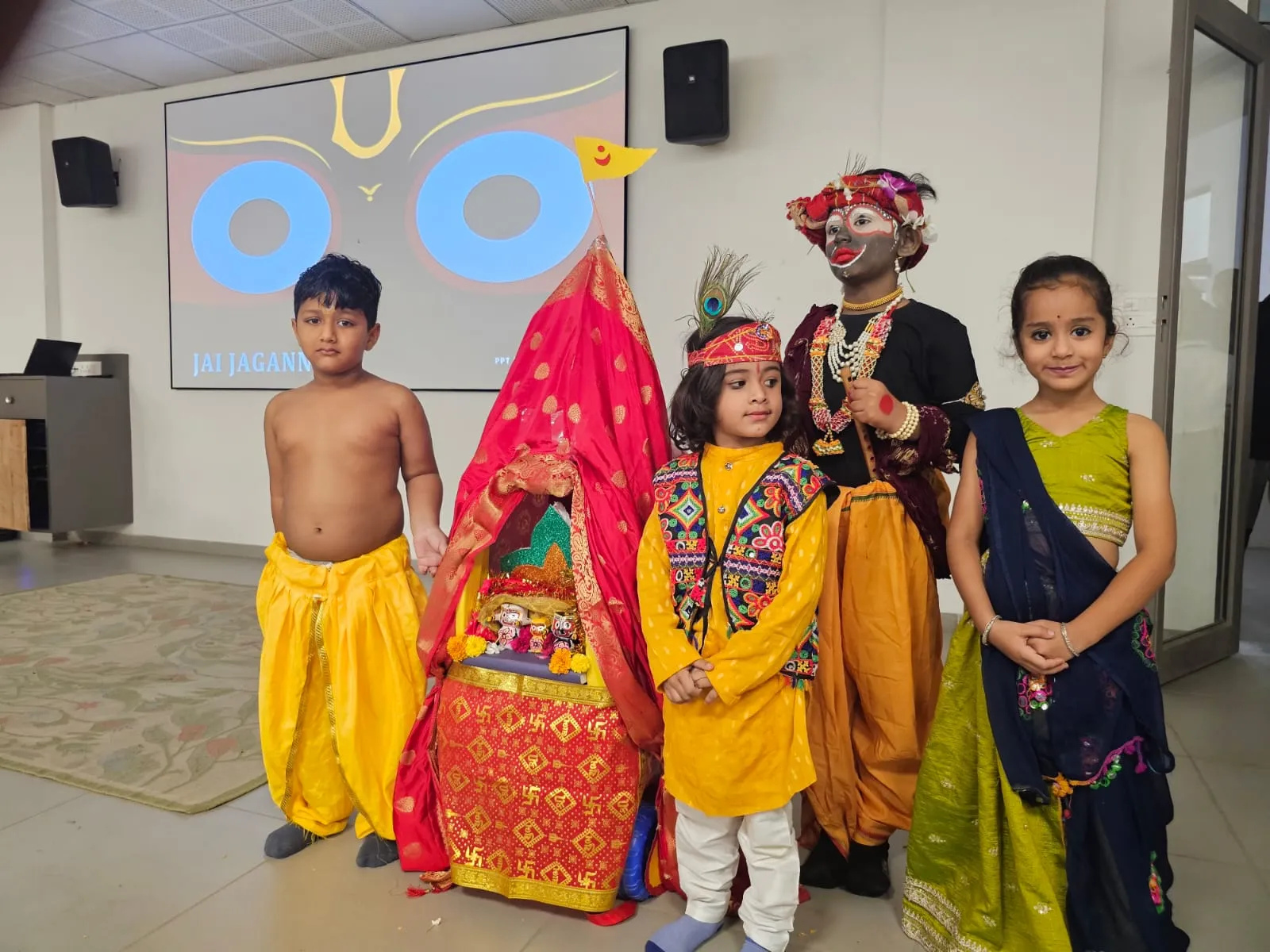 Four young children standing in front of a decorated red and yellow chariot containing Hindu deities during a cultural celebration at Shree Shakti School of Innovation (SSI). The children are wearing traditional Indian clothing, including kurtas, dhotis, and elaborate face paint.