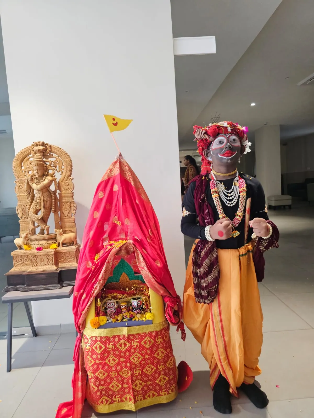 A child wearing traditional cultural attire with striking black and white face paint, a red turban, and pearl necklaces, standing confidently beside a decorated red cloth chariot at Shree Shakti School of Innovation (SSI).