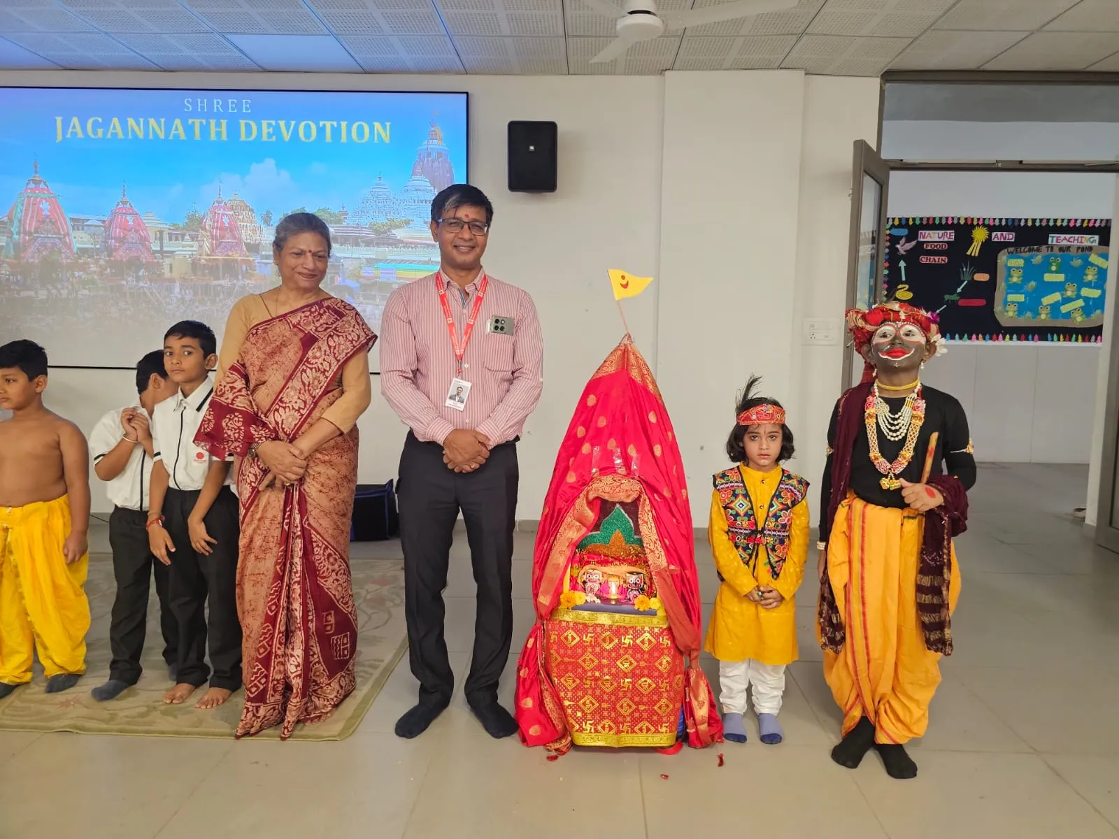 An older woman in a sari and a man standing next to three children in festive attire and a decorated red chariot at Shree Shakti School of Innovation (SSI). Behind them, a projector screen displays "SHREE JAGANNATH DEVOTION.