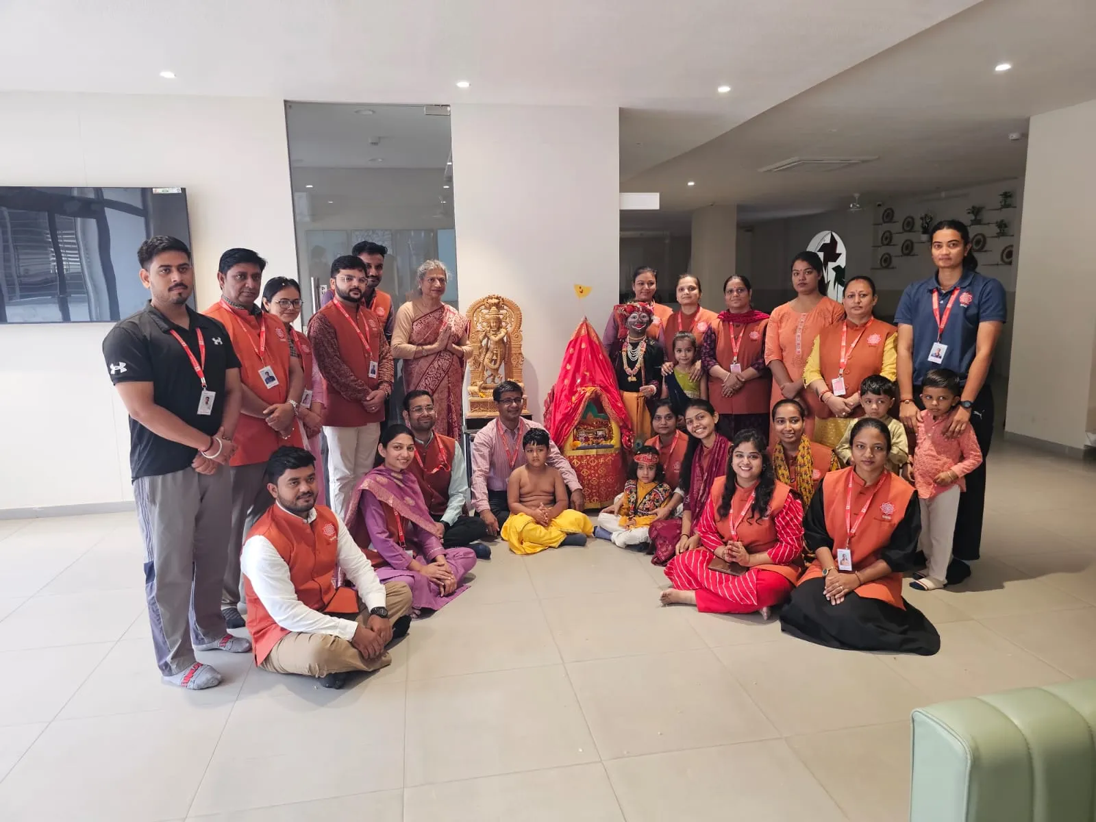 A large group of staff members wearing matching orange lanyards and vests alongside children in traditional dress, gathered around a decorated red chariot for a group picture at Shree Shakti School of Innovation (SSI).