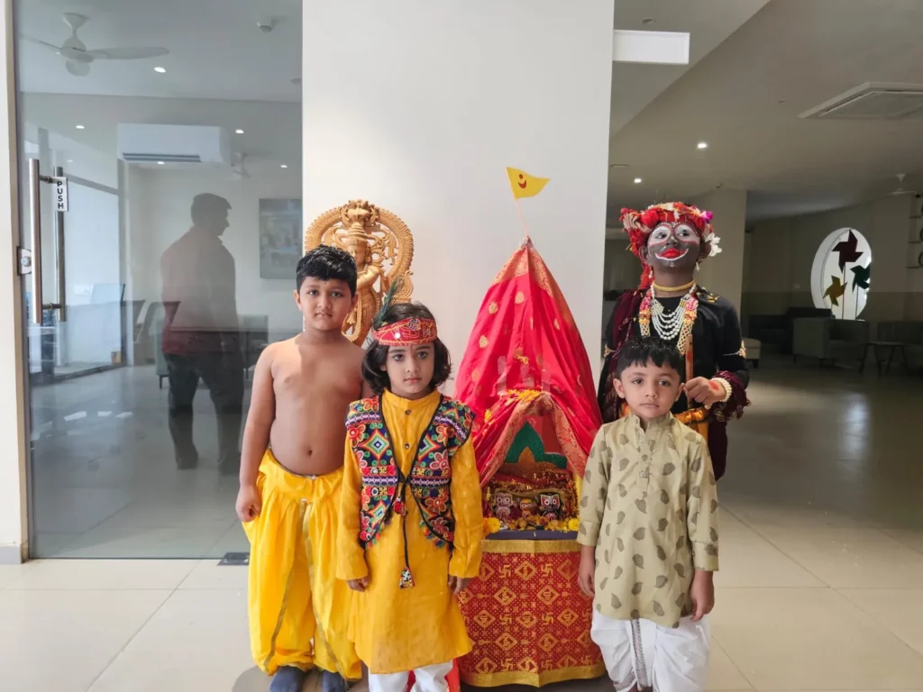 Four young children standing in front of a decorated red and yellow chariot containing Hindu deities during a cultural celebration at Shree Shakti School of Innovation (SSI). The children are wearing traditional Indian clothing, including kurtas, dhotis, and elaborate face paint.