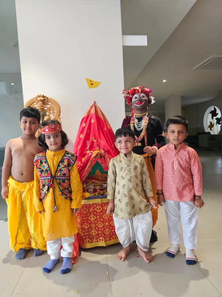 Four young children standing in front of a decorated red and yellow chariot containing Hindu deities during a cultural celebration at Shree Shakti School of Innovation (SSI). The children are wearing traditional Indian clothing, including kurtas, dhotis, and elaborate face paint.