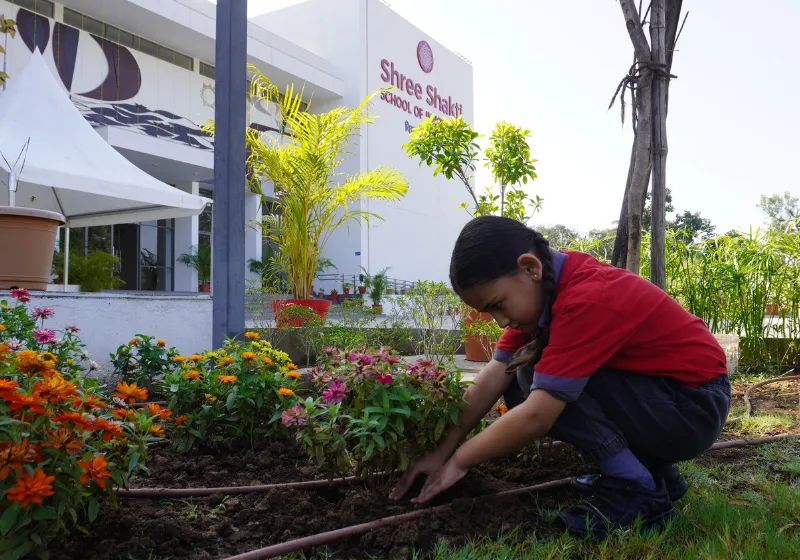 A young female student at SSI Ambaji participating in an outdoor gardening activity, tending to a vibrant flower bed in front of the Shree Shakti school building.