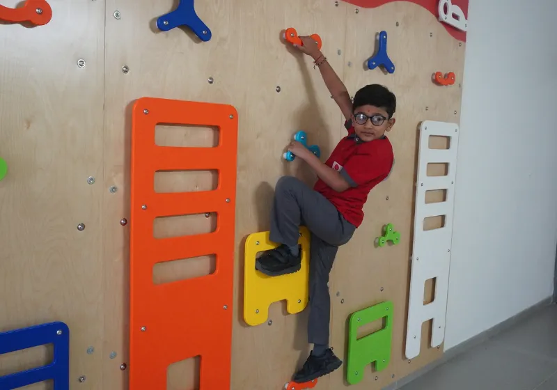 An active student safely navigating the colorful indoor rock climbing wall at SSI Ambaji, an activity designed to promote physical education and motor skills.