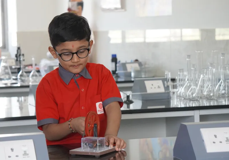 A young student in a red uniform conducting a hands-on physics lab experiment using a mechanical wheel apparatus at the SSI Ambaji science laboratory.