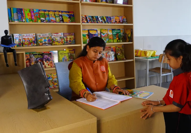 SSI_library_desk_teacher_and_student A teacher at the SSI Ambaji school library desk assisting a student in a red school uniform with book issuing and reading guidance.